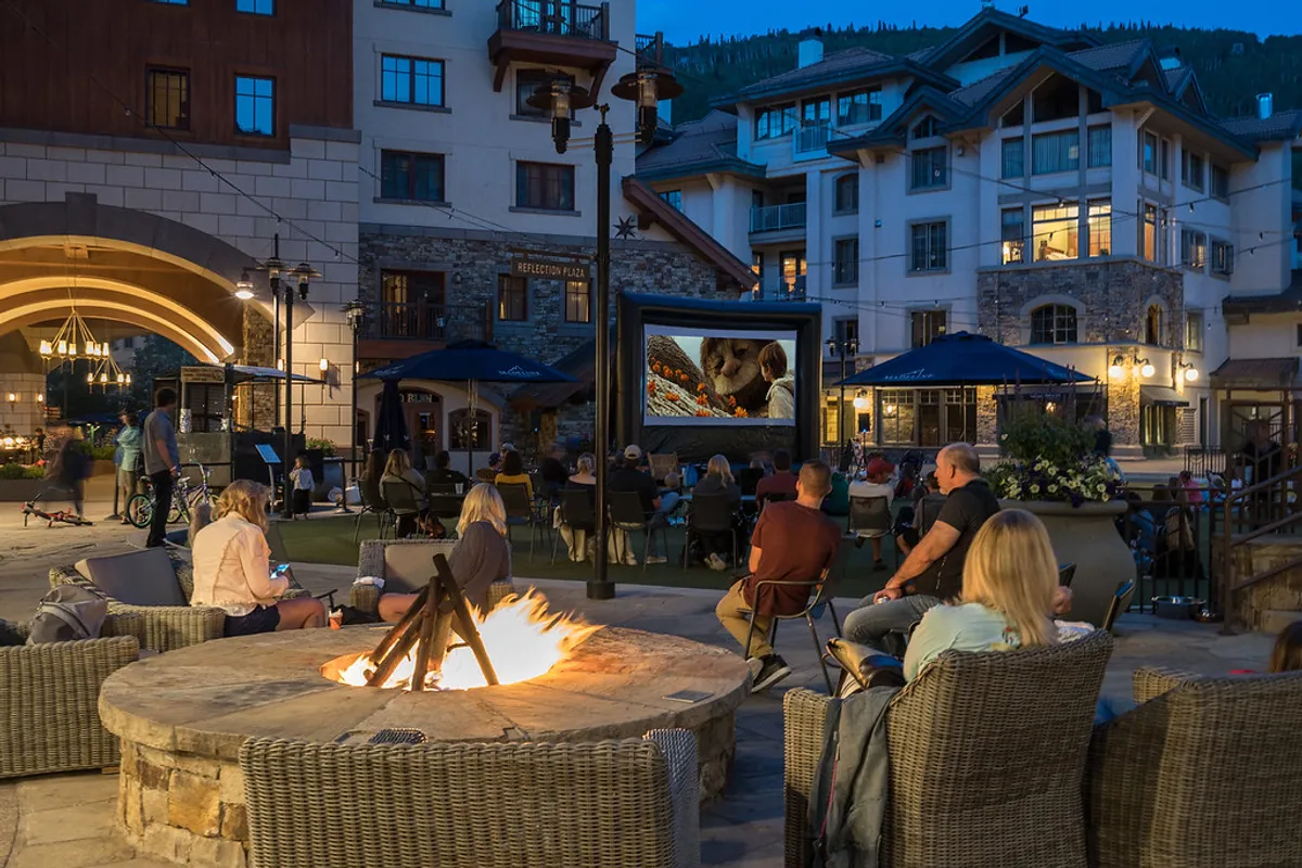 An inviting outdoor communal area with a fire pit, movie screen, comfortable seating, and surrounding architecture. The scene invites group gatherings for movies or conversation under the open sky.