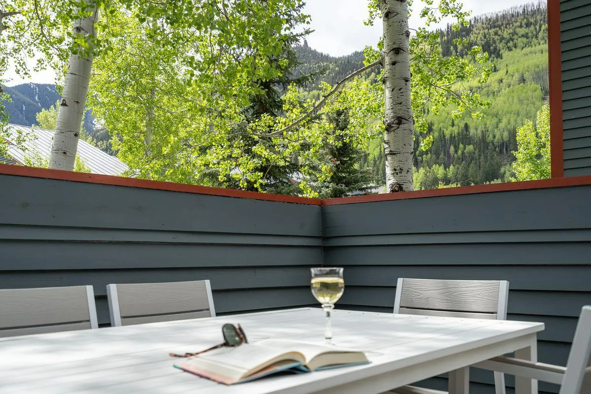 A serene outdoor scene featuring a table set with a book and a glass of wine, nestled among trees. The setup suggests an inviting spot for relaxing, reading, or sharing drinks in a tranquil natural setting.