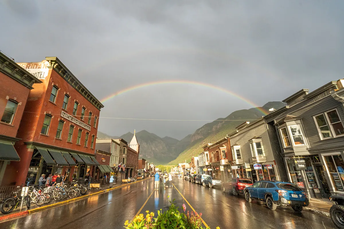 A vibrant street scene in Telluride featuring historic buildings, a colorful rainbow arching over wet streets, bicycles, and mountains in the background. The atmosphere is lively and welcoming, ideal for exploring local culture and dining.