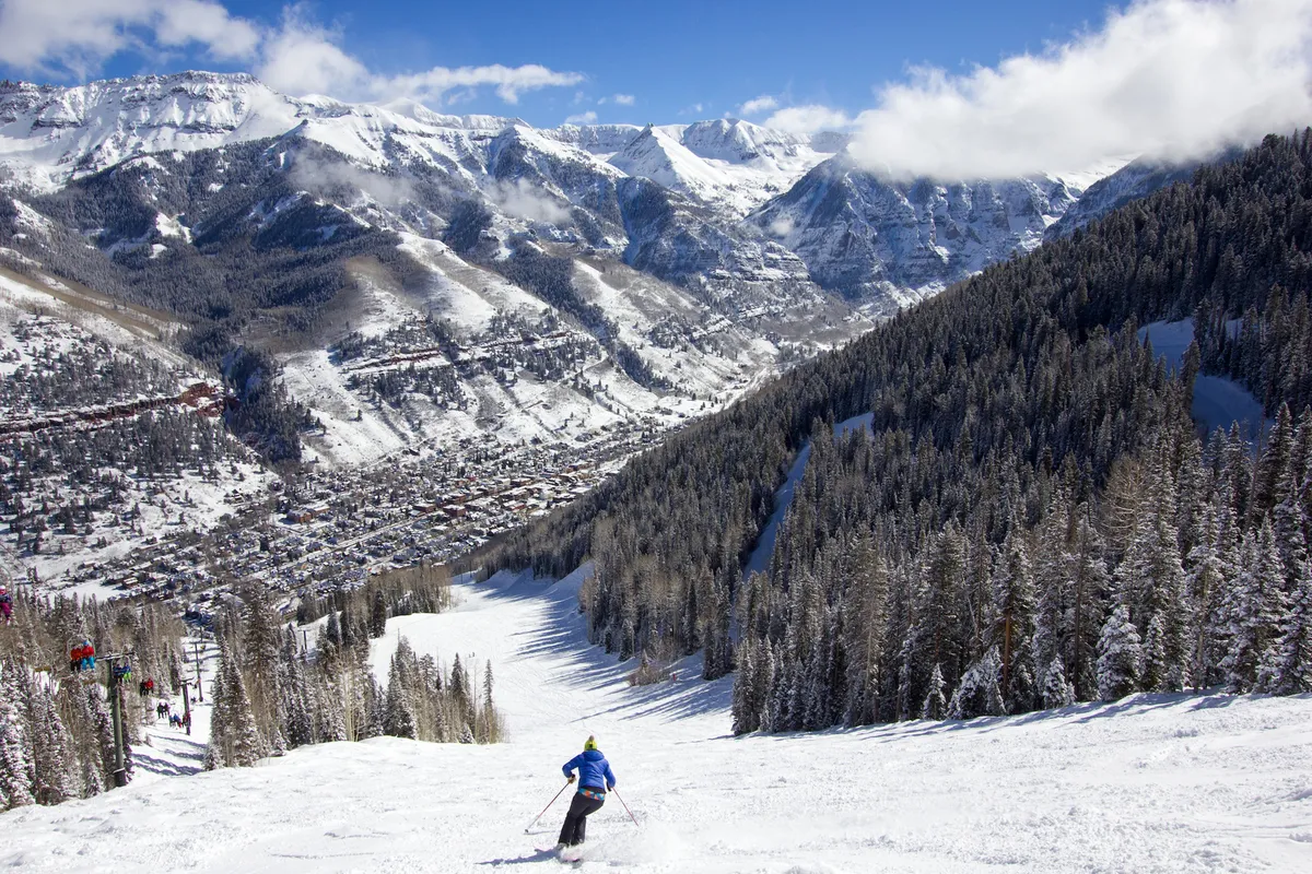 A skier glides down snow-covered slopes with a panoramic view of the mountain range and the charming town below. The bright winter sun glistens off the pristine snow, evoking the excitement and adventure of Telluride's renowned ski season.