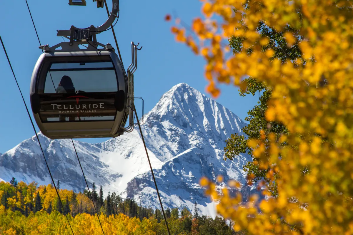 A gondola ascends over a dramatic landscape of snow-capped mountains and bright autumn foliage, providing a panoramic view of the area’s year-round adventure possibilities. The setting blends winter excitement with the promise of summer exploration.