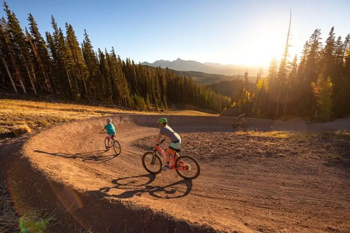 Mountain bikers navigate a winding trail at sunset with expansive mountain views. The scene is energetic, showing families or groups enjoying an active adventure in a beautiful natural setting close to home.