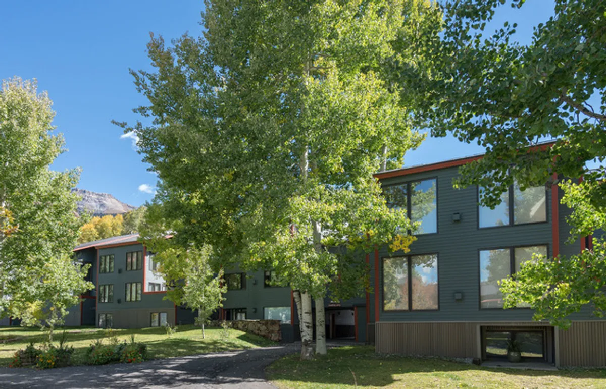 A modern condominium complex set amidst lush green trees, with a dramatic mountain backdrop. The building’s contemporary architecture blends into the natural landscape, and the outdoor area is inviting for walks, play, or relaxation.