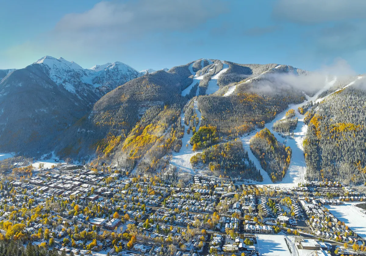 A sweeping exterior view captures snow-capped mountains, active ski slopes, pockets of autumn foliage, and the quaint town below. The scene radiates energy and adventure, inviting viewers to imagine themselves racing down the slopes or exploring the vibrant community nestled in the valley.