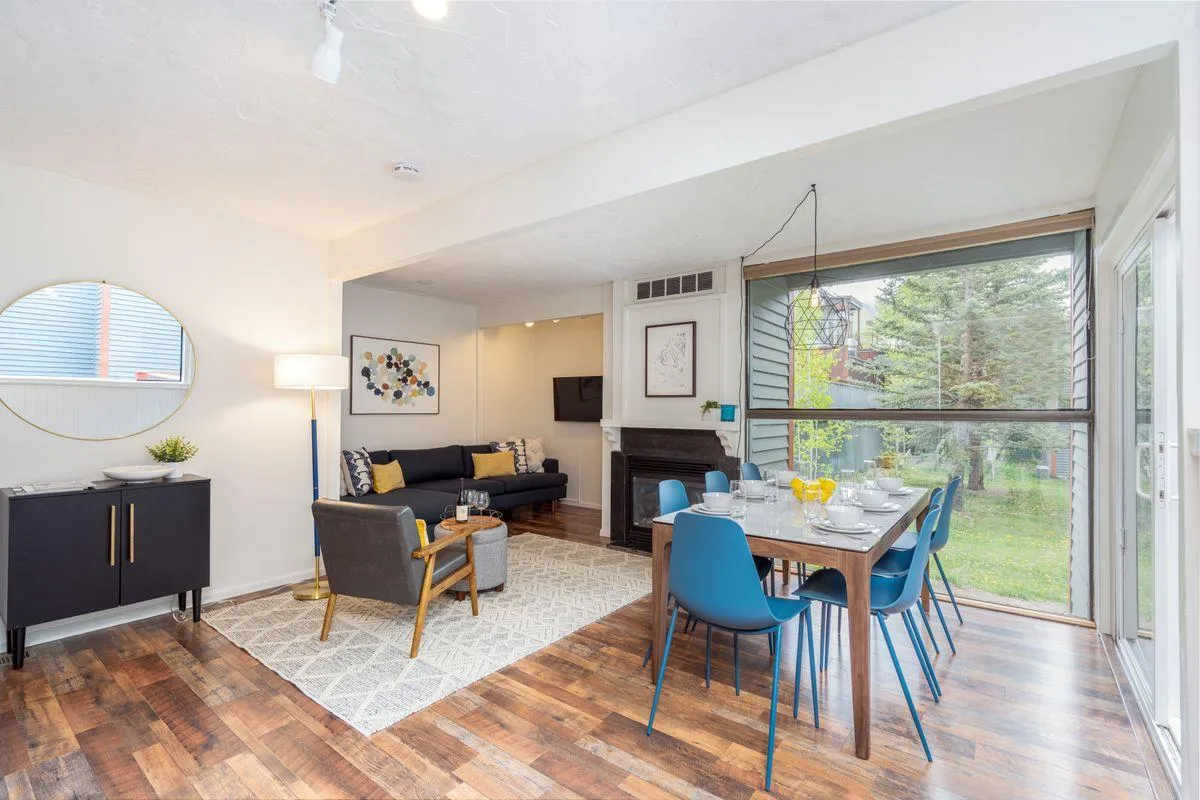 A spacious, welcoming living and dining area featuring a cozy sofa, fireplace, dining table, wall art, and a large window letting in natural light. The arrangement invites both relaxation and group gatherings, with the living and dining spaces seamlessly connected.