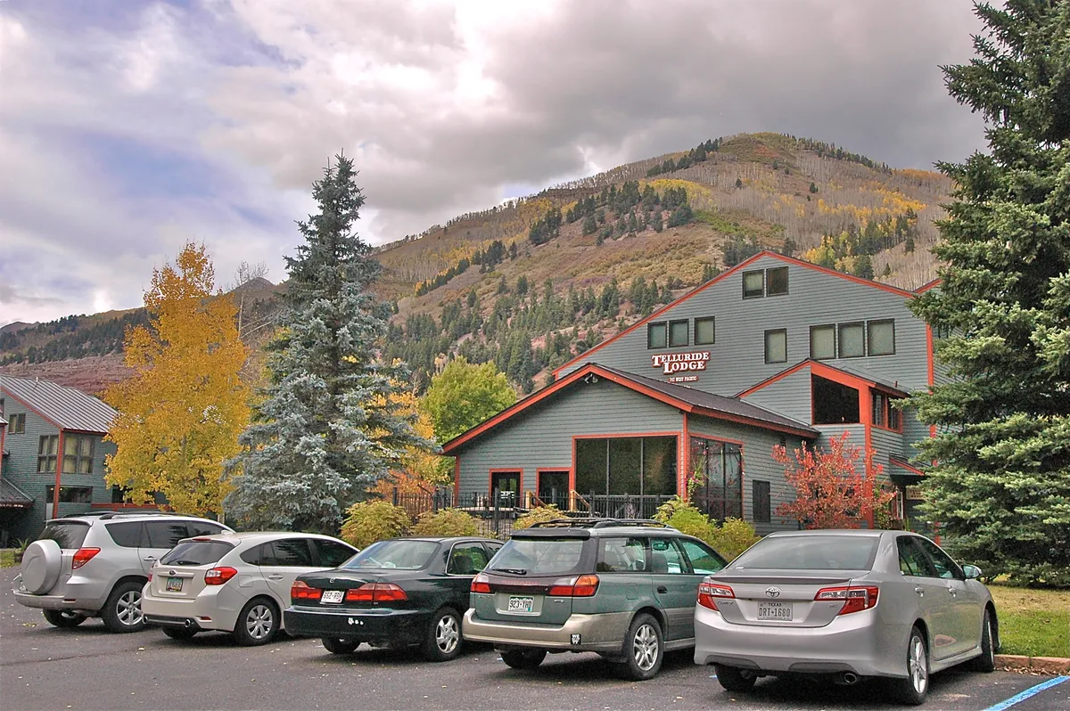 An exterior shot of the lodge building surrounded by trees, with the mountain backdrop and visible parking. The scene feels inviting and hints at the property's welcoming atmosphere, convenient parking, and proximity to nature.