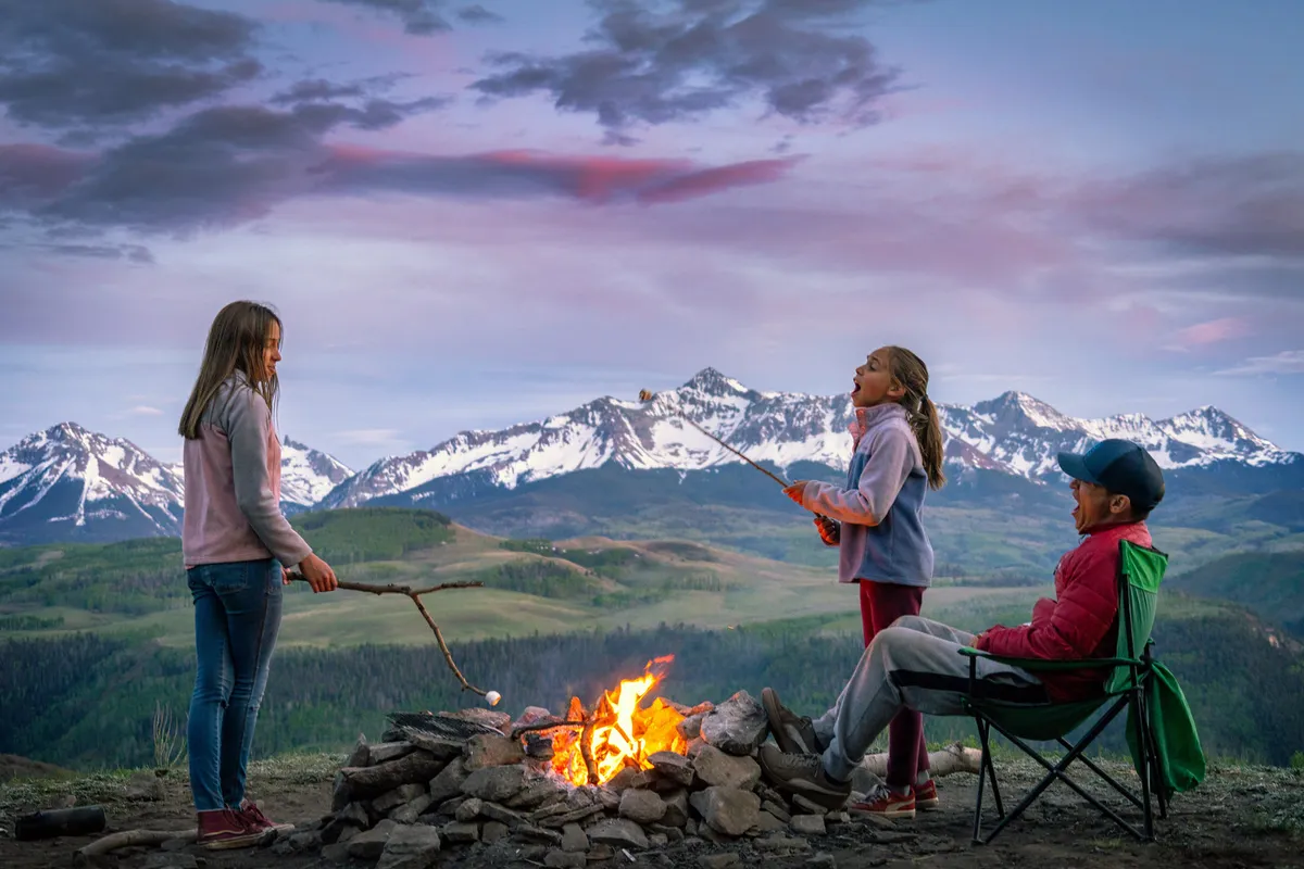 Children roast marshmallows over a campfire with a mountain landscape in the background. The atmosphere is joyful, capturing group fun, storytelling, and togetherness surrounded by nature.