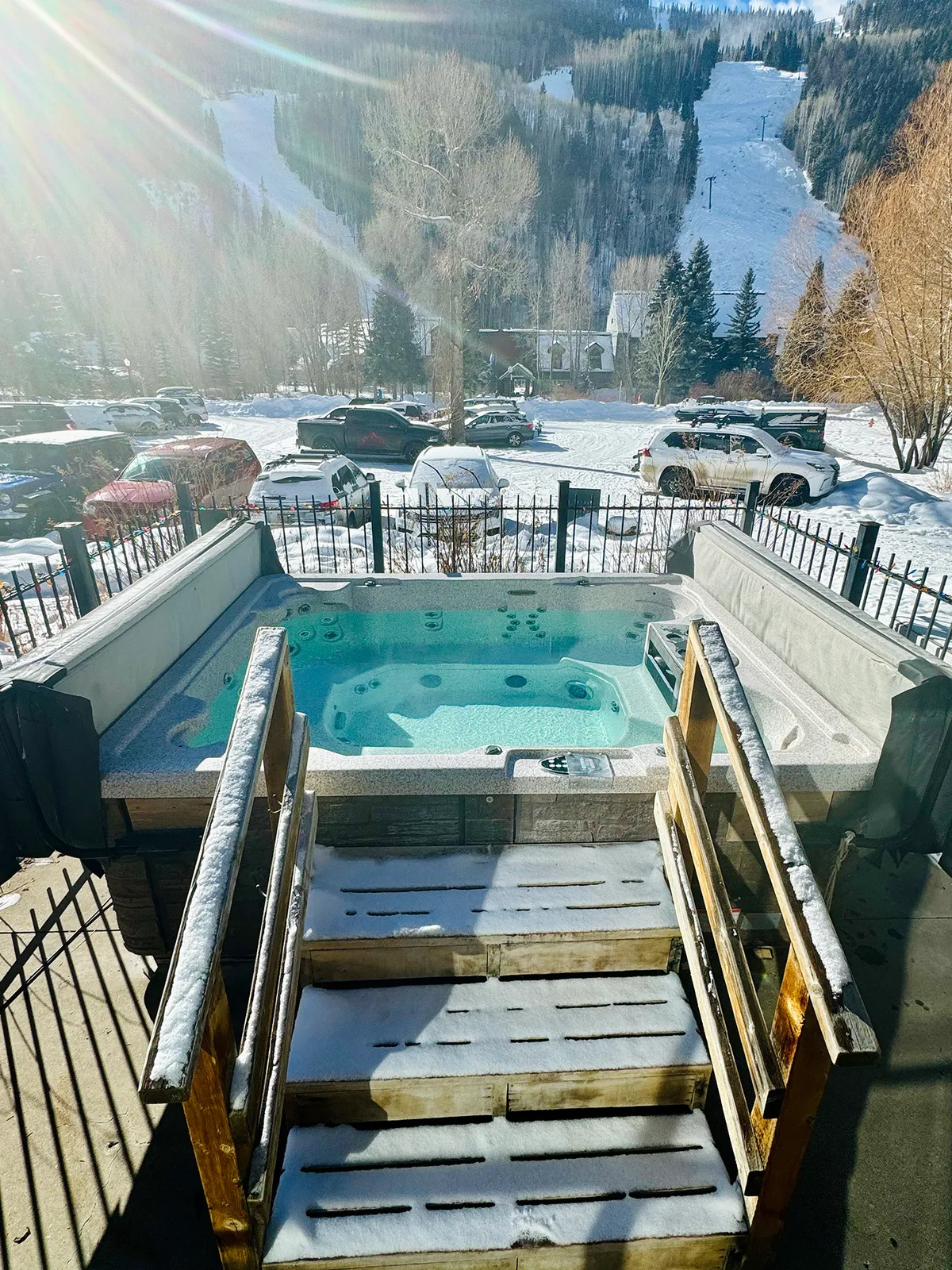 An inviting hot tub sits outside a home, surrounded by snow-covered steps, with a fenced yard and dramatic mountain backdrop. The warm glow from inside contrasts with the cool winter air, creating a cozy, rejuvenating setting ideal for après-ski relaxation or stargazing.