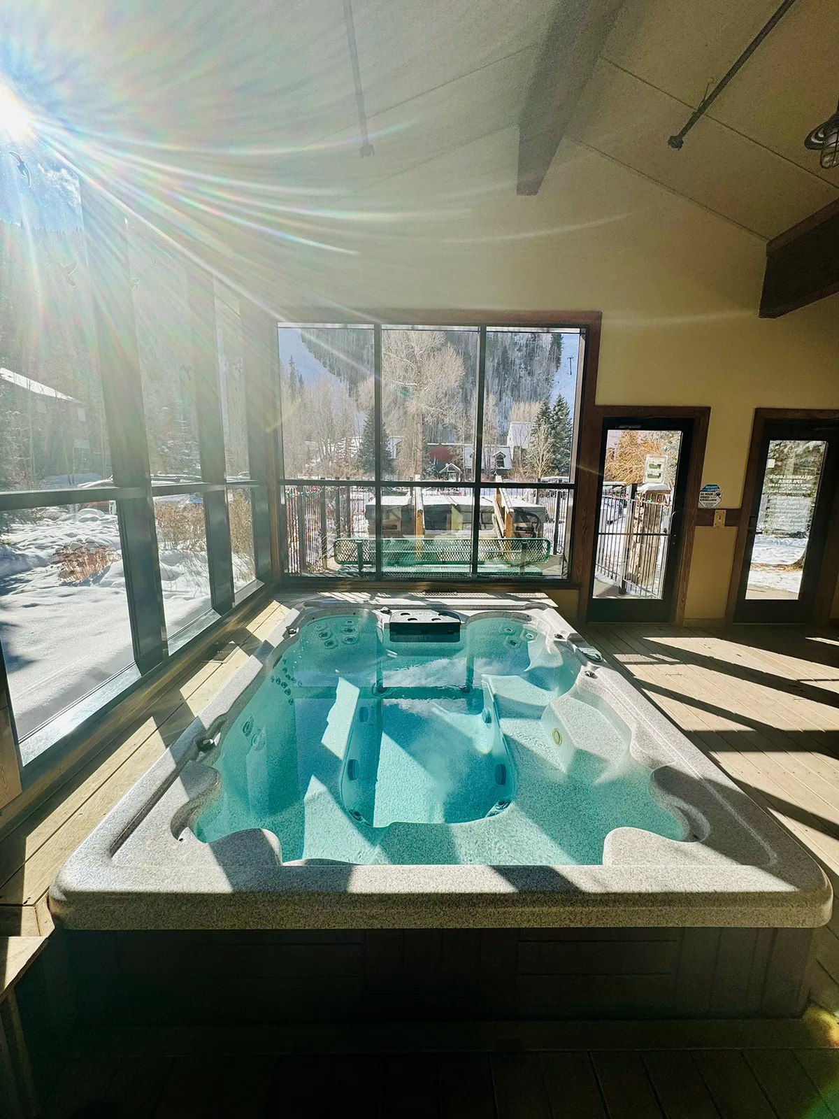 A tranquil interior scene showcases a hot tub set before large windows, through which stunning mountain vistas unfold. The warm, inviting water contrasts with the crisp mountains outside, evoking relaxation after a day of adventure.